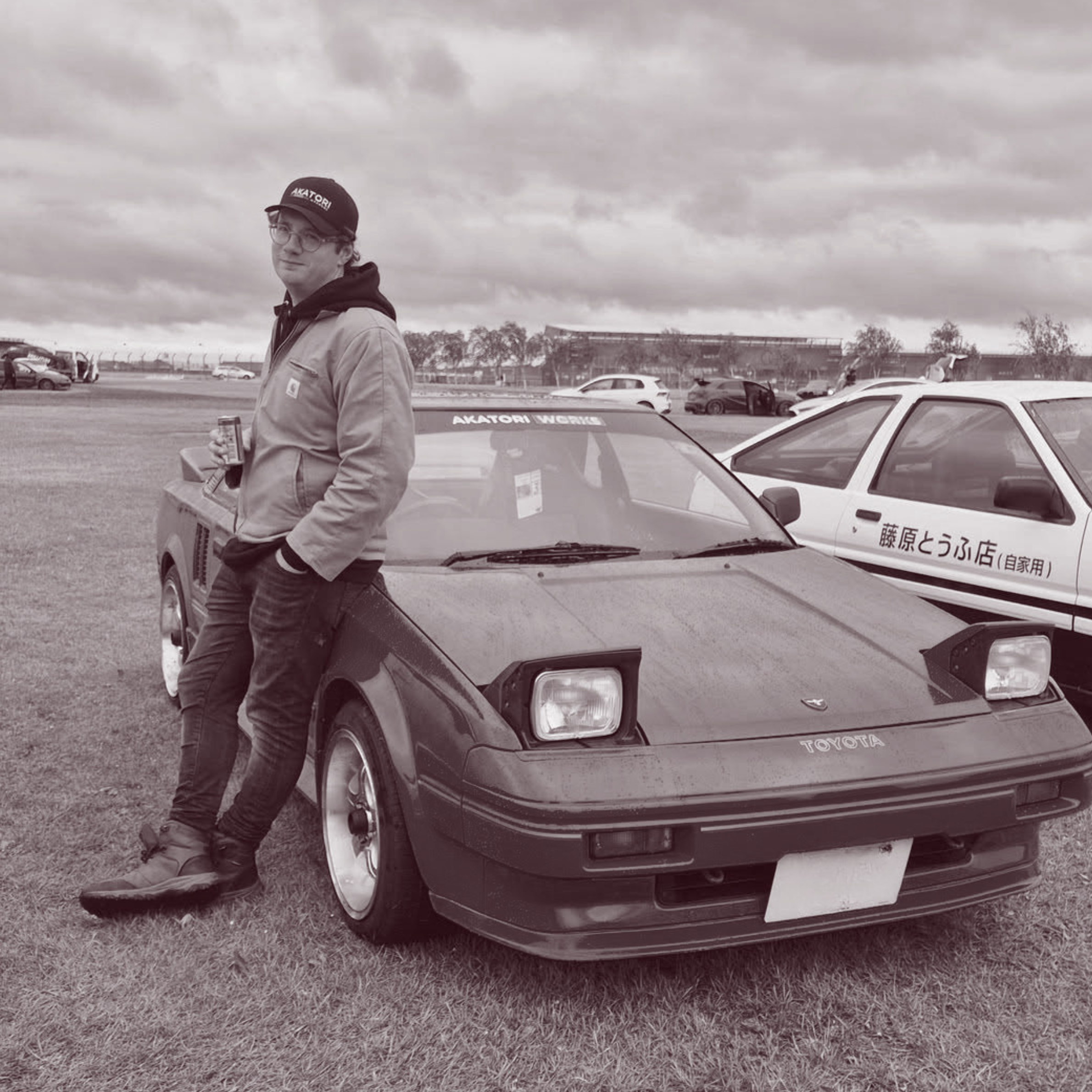 Person standing next to a Toyota car in an outdoor setting with other vehicles and buildings in the background.
